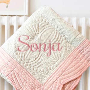 Puede incluir: Una manta acolchada cuadrada que presenta un centro de color crema con intrincados patrones de costura y el nombre Sonja bordado en letra cursiva rosa. La manta est&aacute; enmarcada por un borde ancho de color rosa s&oacute;lido con costuras lineales.