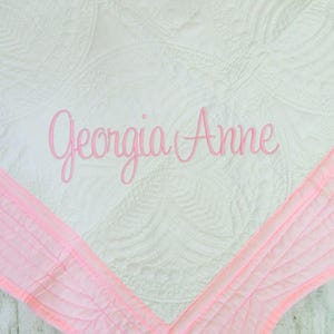 Puede incluir: Una manta blanca acolchada con un patr&oacute;n circular repetitivo y un borde rosa suave. El nombre Georgia Anne est&aacute; bordado en el centro con una fuente cursiva rosa, a&ntilde;adiendo un toque personalizado al dise&ntilde;o de la tela texturizada.