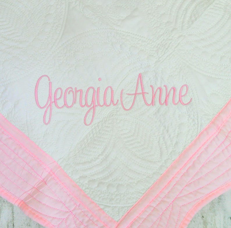 Puede incluir: Una manta blanca acolchada con un patr&oacute;n circular repetitivo y un borde rosa suave. El nombre Georgia Anne est&aacute; bordado en el centro con una fuente cursiva rosa, a&ntilde;adiendo un toque personalizado al dise&ntilde;o de la tela texturizada.