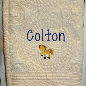 Puede incluir: Una manta acolchada de color crema con intrincados patrones de costura circular. El centro muestra el nombre Colton bordado en hilo azul oscuro, colocado sobre un peque&ntilde;o gr&aacute;fico bordado de un caballo color canela con crines marrones.