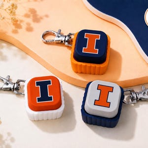 May include: Three square keychains with silver lobster clasps. One has a navy blue top with an orange letter I, one has an orange top with a navy blue letter I, and one has a white top with an orange letter I. Each features a textured base.
