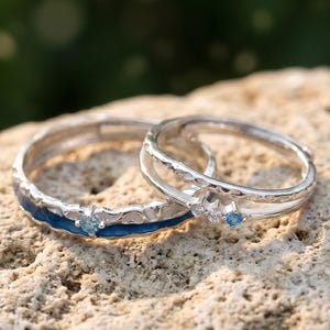 May include: Two silver rings with blue detailing and small blue gemstones. One ring has a blue enamel band, the other a delicate open design. The rings are on a textured, light brown surface, with a blurred green background.
