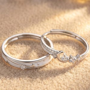 Op de afbeelding: Twee zilveren ringen met hemelse ontwerpen. Eén ring heeft een witte emaille band met ster- en stipdetails en een enkele heldere edelsteen. De andere ring heeft een maan- en sterontwerp met meerdere heldere edelstenen.