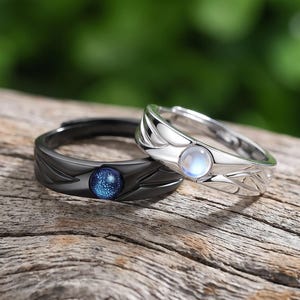Puede incluir: Dos anillos con diseño de banda texturizada. Un anillo es negro mate con una incrustación de piedra circular azul oscuro brillante. El otro anillo es de plata pulida con una incrustación de piedra circular translúcida e iridiscente.