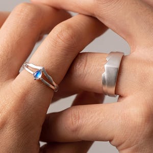 Pode incluir: Um anel de prata com uma pequena pedra da lua azul em forma de lágrima numa banda estilo chevron, emparelhado com uma banda de prata mais larga com acabamento mate e um design geométrico de pico montanhoso no topo.