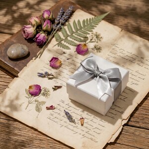 K&ouml;nnte beinhalten: Eine wei&szlig;e Geschenkbox mit silberner Satinschleife liegt auf gealtertem Papier mit handschriftlichem Text. Getrocknete Rosenknospen, Lavendel und ein Farnblatt sind um die Box herum angeordnet.