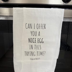 May include: White kitchen towel with black text: "CAN I OFFER YOU A NICE EGG IN THIS TRYING TIME?" and "FRANK REYNOLDS". The towel is hanging on an oven door, a kitchen appliance.