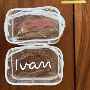 May include: Two rectangular clear plastic toiletry bags with white zipper trim. The top bag features the name Carmella in pink script lettering. The bottom bag features the name Ivan in white script lettering. Both bags have a transparent design.
