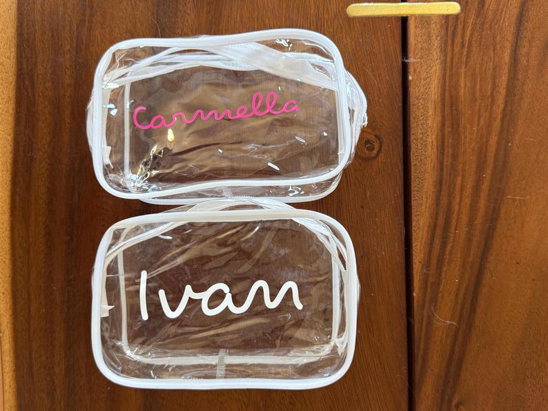 May include: Two rectangular clear plastic toiletry bags with white zipper trim. The top bag features the name Carmella in pink script lettering. The bottom bag features the name Ivan in white script lettering. Both bags have a transparent design.