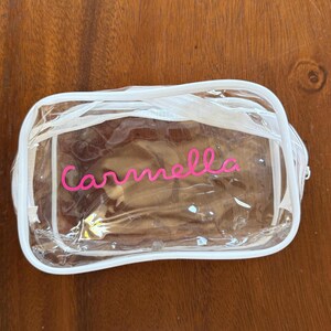 May include: A rectangular clear plastic pouch with white trim and a white zipper closure. The name Carmella is printed in a bright pink cursive font on the front of the transparent bag, which features rounded corners and a structured design.