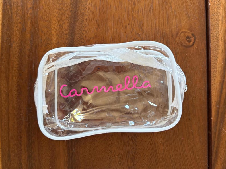 May include: A rectangular clear plastic pouch with white trim and a white zipper closure. The name Carmella is printed in a bright pink cursive font on the front of the transparent bag, which features rounded corners and a structured design.