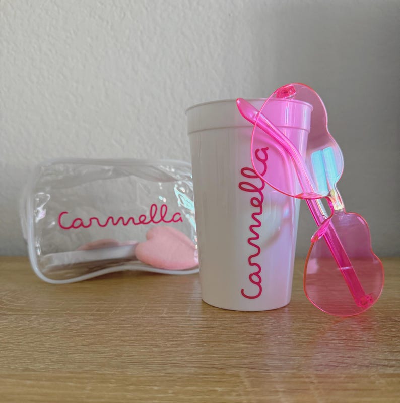 May include: A white plastic cup and a clear zippered pouch, both featuring the name Carmella in pink script. A pair of translucent pink heart-shaped sunglasses rests against the side of the cup, which also contains a small pink heart-shaped object.