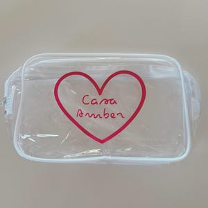 May include: A rectangular clear plastic pouch with white trim and a white zipper closure. The front features a large red heart outline with the text Casa Amber written in a cursive red font inside the center of the heart shape.