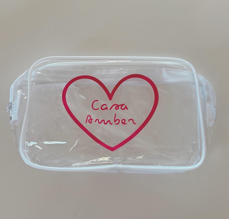 May include: A rectangular clear plastic pouch with white trim and a white zipper closure. The front features a large red heart outline with the text Casa Amber written in a cursive red font inside the center of the heart shape.