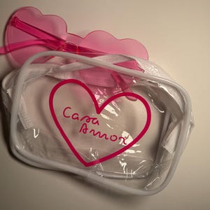 May include: A clear rectangular zippered pouch with white trim and a pink heart graphic containing the text Casa Amor, paired with translucent pink heart-shaped sunglasses resting on top of the pouch.