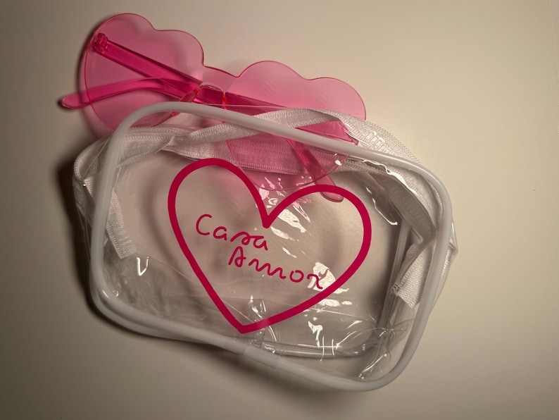 May include: A clear rectangular zippered pouch with white trim and a pink heart graphic containing the text Casa Amor, paired with translucent pink heart-shaped sunglasses resting on top of the pouch.