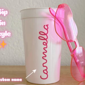 May include: A white plastic cup featuring the name Carmella in pink script lettering, accompanied by a pair of translucent pink heart-shaped sunglasses. The text Sip in style and custom name with an arrow are visible on the left side of the cup.