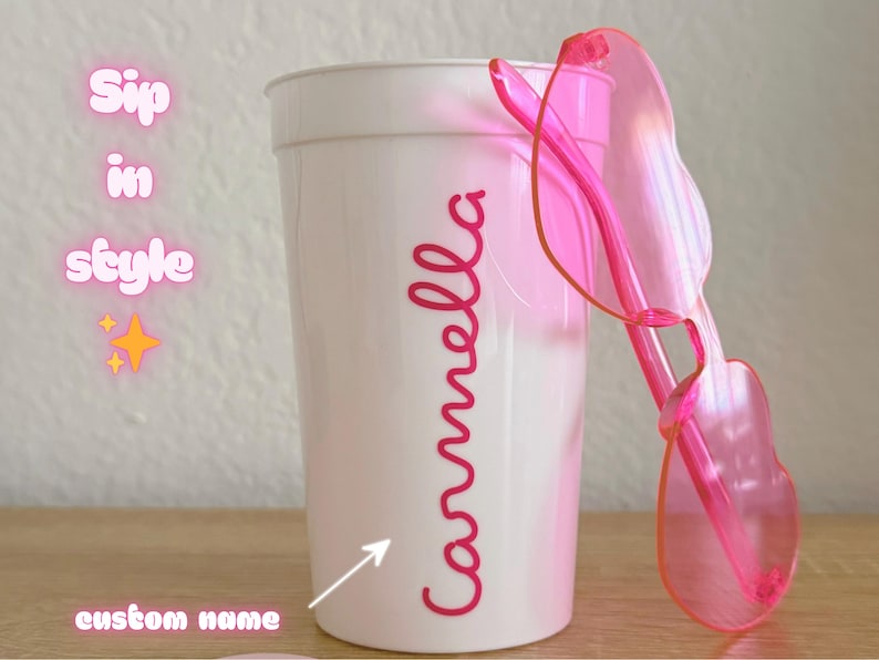 May include: A white plastic cup featuring the name Carmella in pink script lettering, accompanied by a pair of translucent pink heart-shaped sunglasses. The text Sip in style and custom name with an arrow are visible on the left side of the cup.