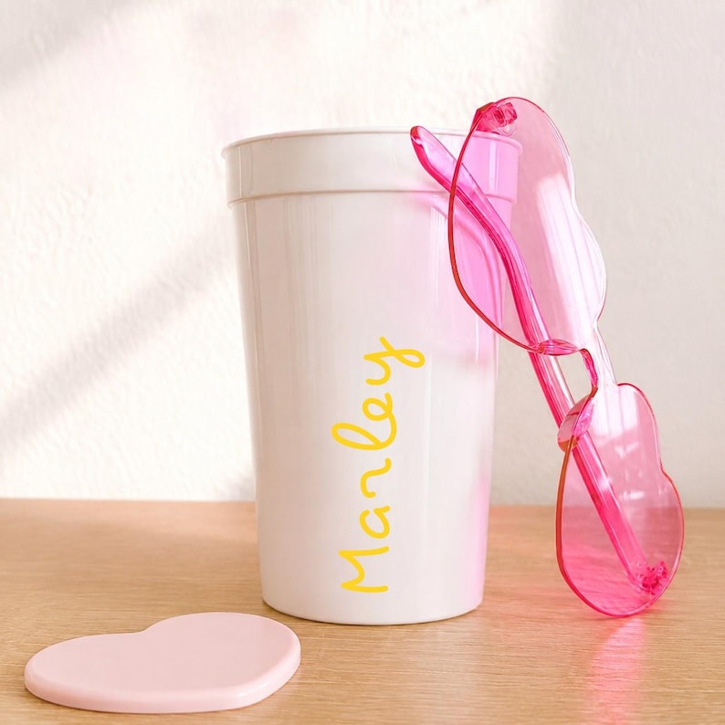May include: A white plastic cup with the name Marley printed in yellow script font. Leaning against the cup are translucent pink heart-shaped sunglasses. A small, light pink heart-shaped object lies flat on the surface in front of the cup.