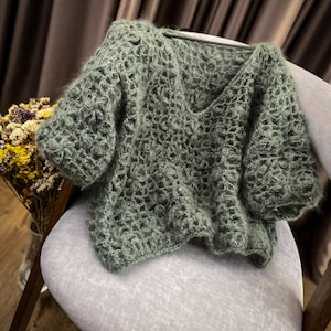 Può includere: Un maglione verde salvia a maniche corte e scollo a V, con trama aperta. Il maglione è realizzato in un filato morbido e strutturato ed è esposto su una sedia grigia. Il design a trama aperta crea un aspetto delicato e arioso.