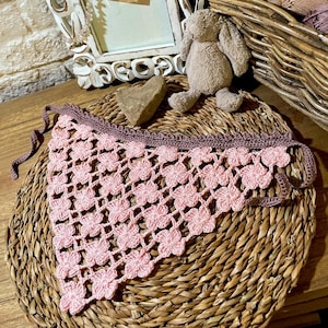 Könnte beinhalten: Ein handgefertigtes, rosa gehäkeltes Bandana mit floralem Muster. Das Bandana hat einen dunkelmalvenfarbenen Rand und Bindebänder. Das Bandana wird auf einer gewebten, kreisförmigen Oberfläche präsentiert.
