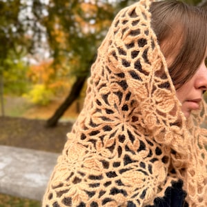 Può includere: Uno scialle all'uncinetto beige con un motivo floreale. Il design traforato presenta motivi ripetuti di fiori e foglie. Lo scialle è drappeggiato sulle spalle, con i dettagli intricati del lavoro all'uncinetto ben visibili.