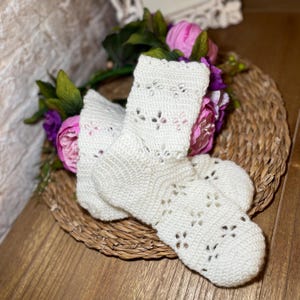 May include: A pair of off-white, crocheted socks with a floral pattern. The socks are displayed on a woven basket, with pink and purple flowers in the background. The socks have a textured appearance and a delicate, openwork design.