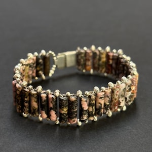 May include: A bracelet made of rectangular, pink and black stones. The stones are strung together with silver-coloured metal beads and wire. The bracelet has a silver-coloured clasp.