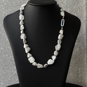 Peut inclure: Un collier composé de pierres irrégulières, gris clair. Les pierres sont enfilées ensemble, formant un collier présenté sur un mannequin noir. Le collier est fermé par un fermoir argenté.