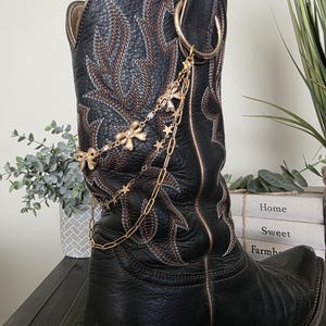 May include: A black leather cowboy boot featuring intricate brown stitching and a gold-toned metal chain accessory. The decorative chain is attached to a large ring and includes small gold bows and star-shaped charms draped across the side.