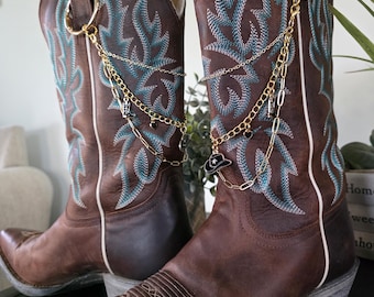 May include: A pair of brown leather cowboy boots featuring teal embroidery and white piping. Each boot is adorned with a gold-toned metal chain accessory that attaches to a ring, featuring small charms shaped like cowboy hats and boots.