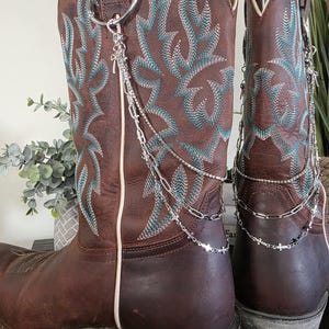May include: A pair of brown leather cowboy boots featuring teal embroidered patterns on the shaft. Each boot is adorned with a silver-toned metal chain accessory that attaches to a ring, featuring delicate cross charms and a layered link design.