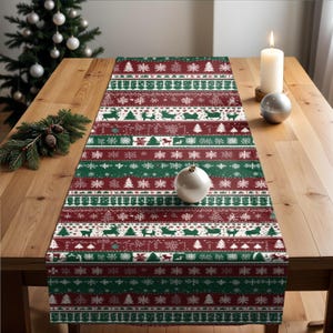 May include: A Christmas-themed table runner with a festive pattern of reindeer, trees, and snowflakes in red, green, and white. The runner is displayed on a wooden table, with a candle and ornaments nearby.