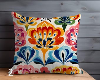 May include: Square decorative pillow with a vibrant floral pattern. The design features large, stylized flowers in shades of orange, red, blue, pink, and green against a white background. The pillow is placed on a wooden surface.