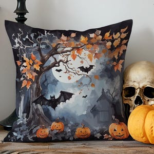 Op de afbeelding: Een decoratief Halloween-kussen met een aquarelillustratie van een spookachtige scène. Het ontwerp toont vleermuizen, pompoenen, een boom met oranje bladeren en een volle maan. Een schedel en een pompoen staan naast het kussen.
