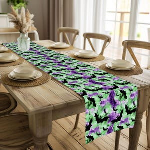 May include: A Halloween-themed table runner featuring a repeating pattern of black bats against a backdrop of green and purple watercolor-style splashes. The runner is displayed on a wooden dining table, adding a festive touch.