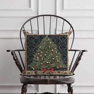 May include: A square cushion with a Christmas tree design. The tree is green with white and gold baubles and a red bow. The cushion has a dark blue background with a floral border, resting on a wooden chair.