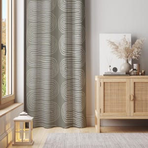 Puede incluir: Una cortina verde salvia con un patrón repetitivo de líneas onduladas blancas. La cortina está junto a una ventana y un mueble de madera con frente tejido. Una linterna blanca está en el suelo frente a la ventana.