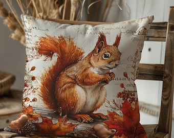 Könnte beinhalten: Quadratisches Zierkissen mit einer detaillierten Illustration eines Eichhörnchens, das eine Nuss hält. Das Eichhörnchen hat rotbraunes Fell und ist von Herbstblättern und Beeren umgeben. Der Hintergrund hat verblasste Schrift.