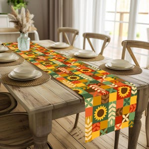 Puede incluir: Un camino de mesa colorido con un diseño de patchwork con girasoles, hojas y formas geométricas en tonos amarillos, naranjas, verdes y rojos. El camino de mesa está colocado sobre una mesa de madera.