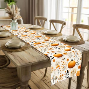Könnte beinhalten: Ein herbstliches Tischläufer-Design mit einem sich wiederholenden Muster aus orangefarbenen Kürbissen, Blumen und Blättern auf cremefarbenem Hintergrund. Der Läufer liegt auf einem Holztisch, der für ein Essen gedeckt ist.