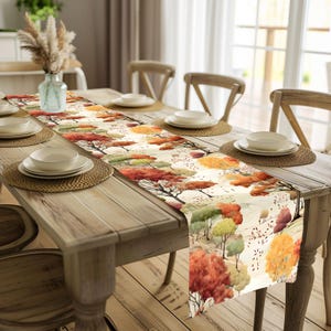 Pode incluir: Um caminho de mesa retangular com um design em aquarela de árvores de outono em tons de vermelho, laranja e verde. O caminho de mesa está colocado sobre uma mesa de madeira montada para uma refeição, com pratos e individuais.