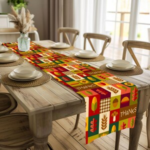 Può includere: Una colorata tovaglia da tavolo del Ringraziamento con un design patchwork. La tovaglia presenta quadrati con illustrazioni a tema autunnale come foglie, girasoli e ghiande. La parola "THANKS" è stampata su diversi quadrati. È posizionata su un tavolo di legno.