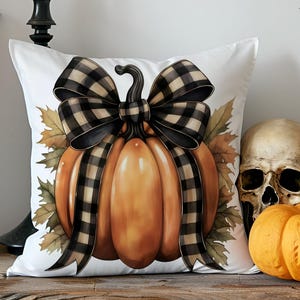 Peut inclure: Un coussin blanc décoratif avec un motif de citrouille et un nœud à carreaux noir et beige. La citrouille est entourée de feuilles d'automne. Une citrouille orange et un crâne sont à côté du coussin.