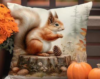 Könnte beinhalten: Ein dekoratives Kissen mit einer detaillierten Illustration eines Eichhörnchens auf einem Baumstumpf mit einem Tannenzapfen. Das Kissen hat einen weichen, cremefarbenen Hintergrund mit Herbstlaub und Kürbissen. Das Eichhörnchen ist braun und weiß.