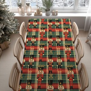 Peut inclure: Une nappe de Noël rectangulaire avec un motif à carreaux rouge, vert et or. La nappe présente des images répétées de casse-noisettes jouets de différentes couleurs. La table est dressée avec des chaises en bois, créant une scène de vacances festive.
