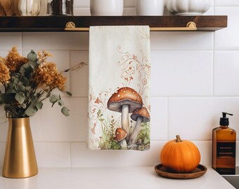 Op de afbeelding: Een crèmekleurige keukendoek met een aquarelillustratie van paddenstoelen. De paddenstoelen hebben bruine doppen met witte stippen en zijn omgeven door groen gras en bladeren. De handdoek hangt aan een donkerbruin houten plankje.