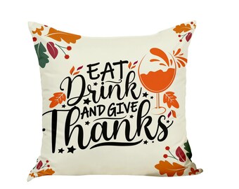 Peut inclure: Coussin carré crème avec les mots "EAT Drink AND GIVE Thanks" en écriture noire. Un verre de vin orange est à droite. Le coussin est décoré de feuilles d'automne orange, rouges et vertes.