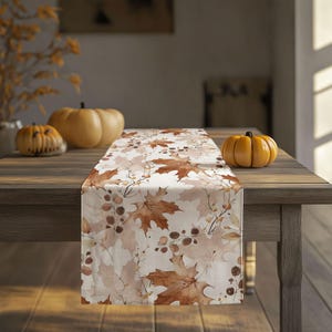 May include: A rectangular table runner with a white background and a repeating pattern of brown autumn leaves and berries. The runner is placed on a wooden table, with pumpkins in the background, creating a fall theme.