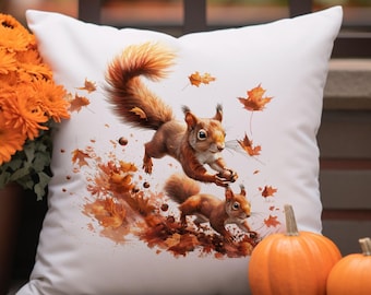 Könnte beinhalten: Quadratisches weißes Kissen mit einer Aquarellillustration von zwei Eichhörnchen, die durch fallende Herbstblätter rennen. Die Eichhörnchen sind braun mit buschigen Schwänzen, und die Blätter sind orange und braun. Zwei kleine Kürbisse sind im Vordergrund.